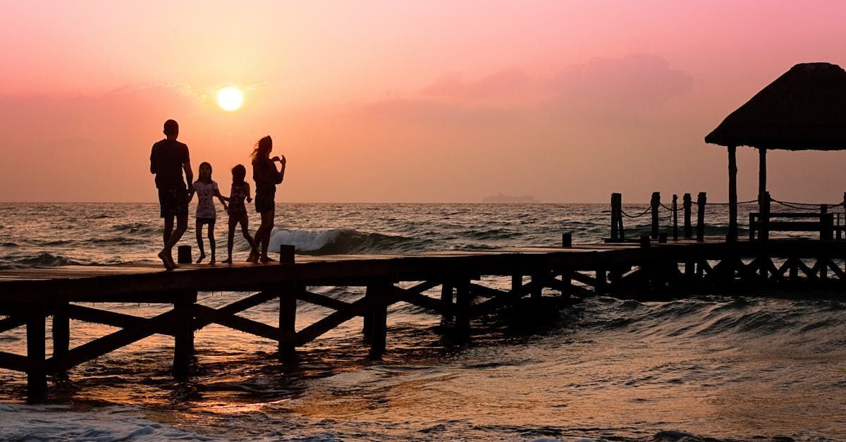 Family on a pier at sunset, silhouetted against orange sky. Waves lap below.