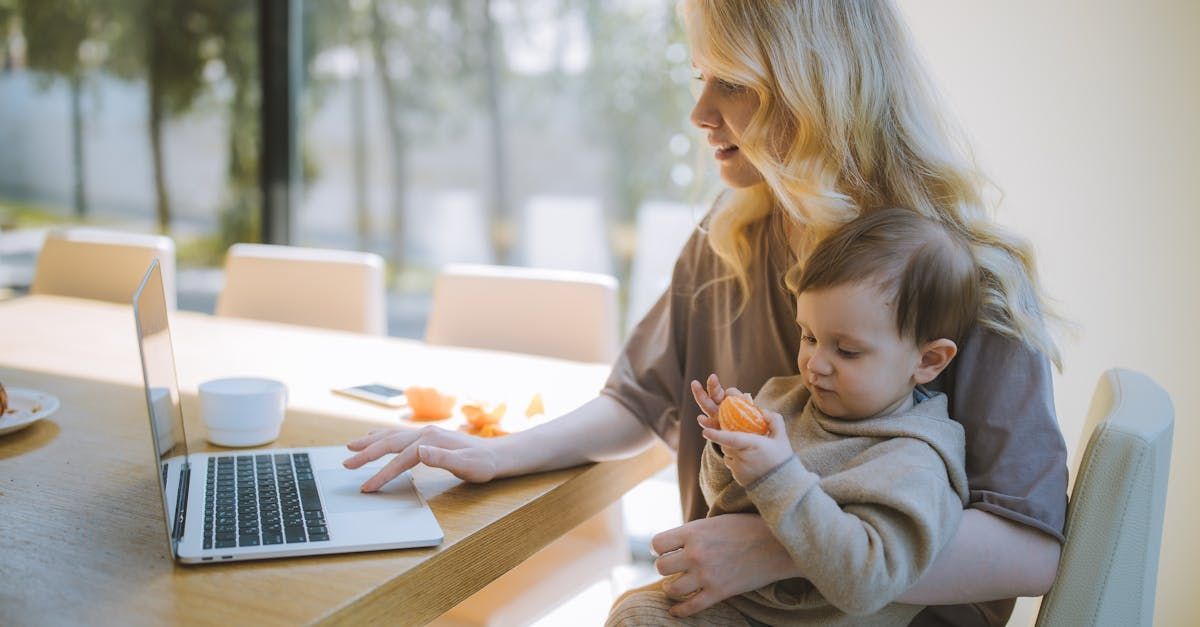 Blonde woman works on laptop at table, holding baby eating a snack indoors.