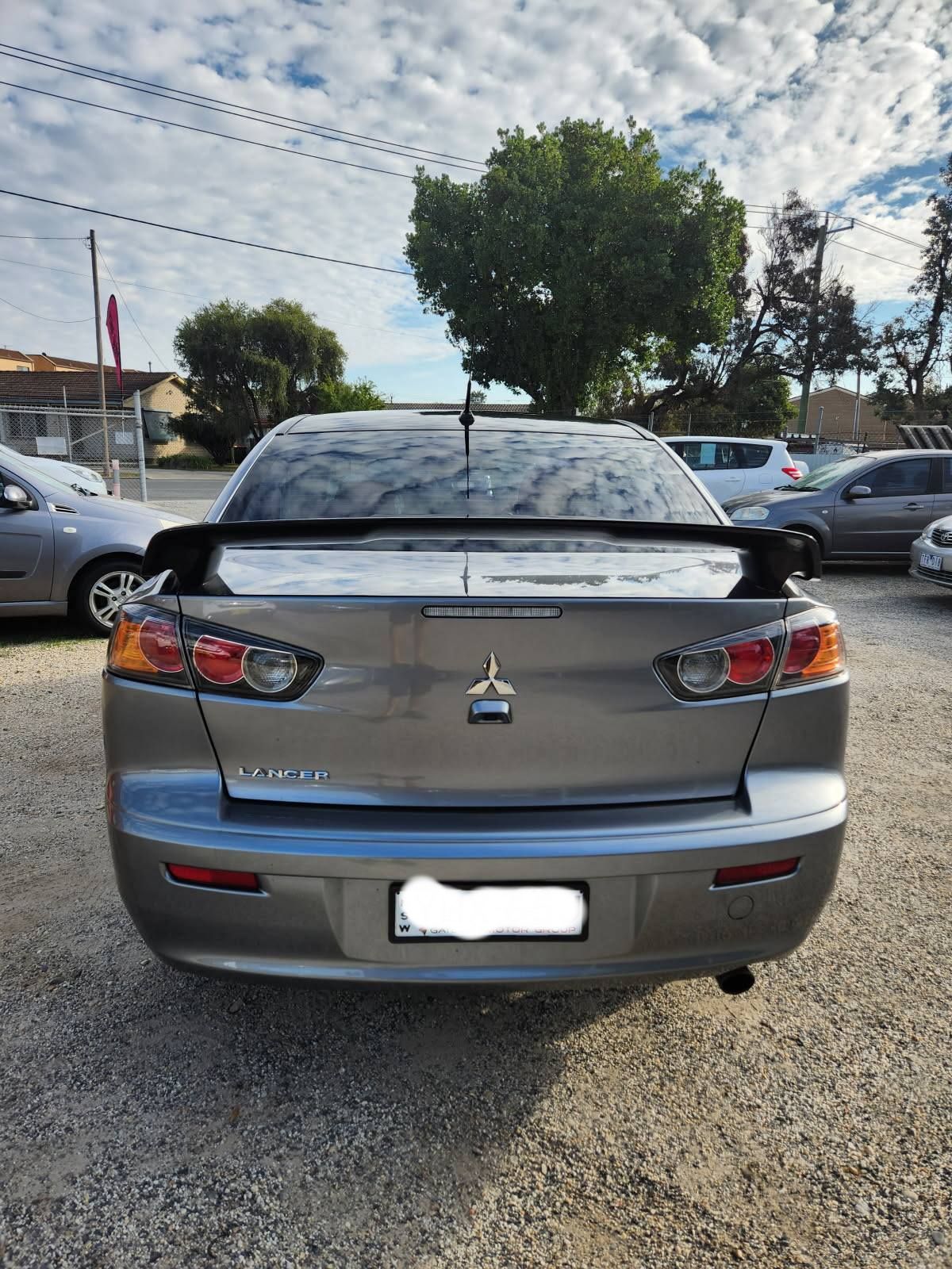 A gray Mitsubishi Lancer from behind, parked outside on a sunny day. The license plate reads YHX-65J.