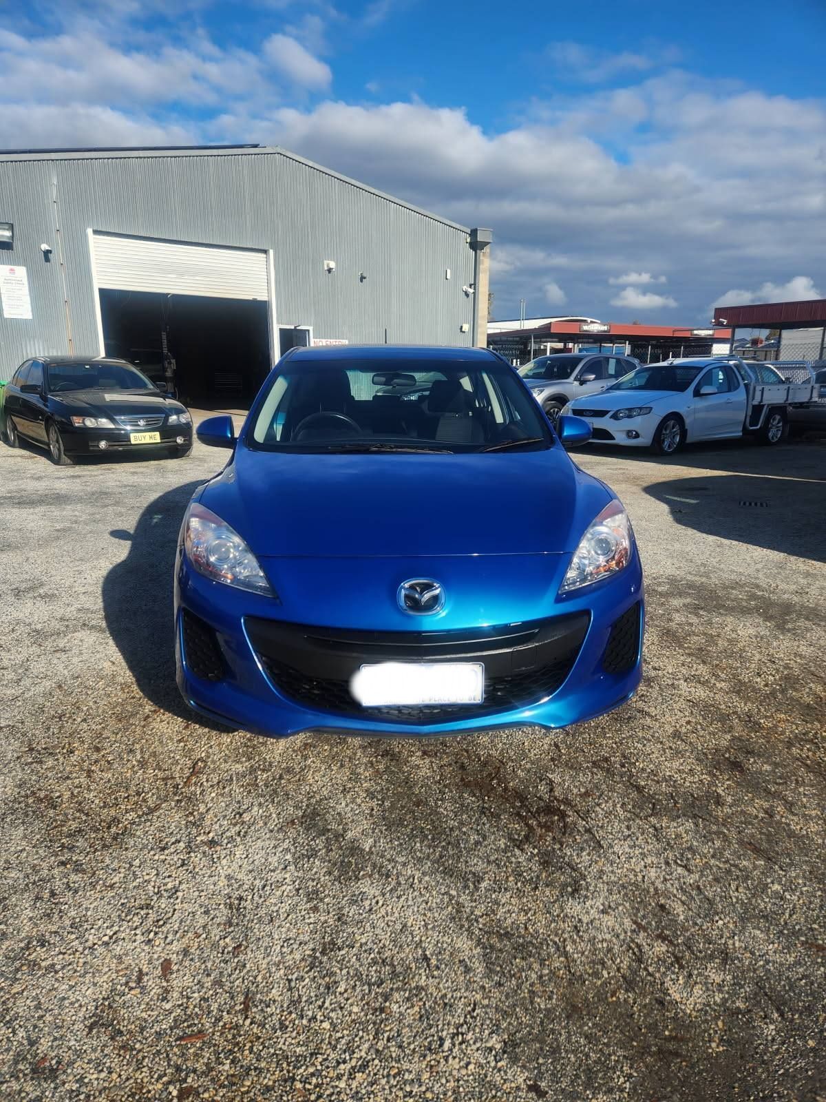 Blue Mazda 3 hatchback parked on gravel in front of a metal building on a sunny day.