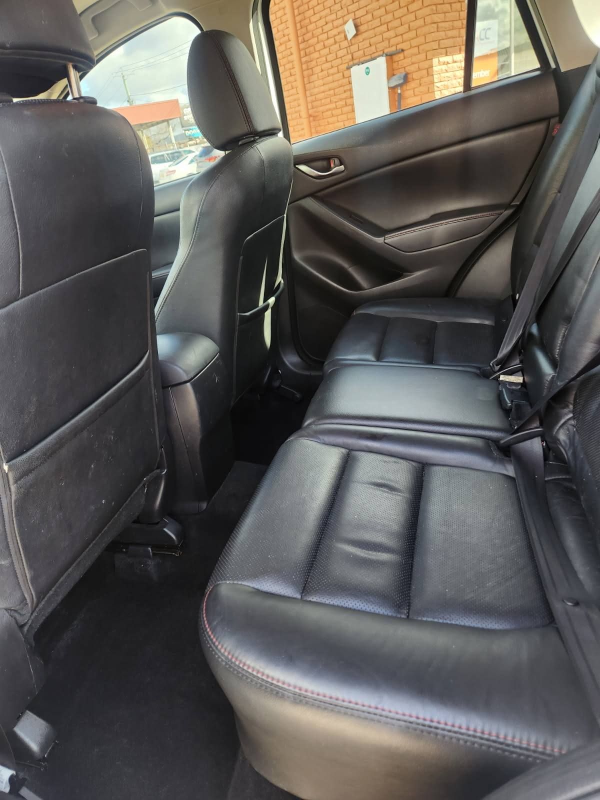 Black leather back seats of a vehicle with a center armrest. Interior view with windows and outside brick wall visible.