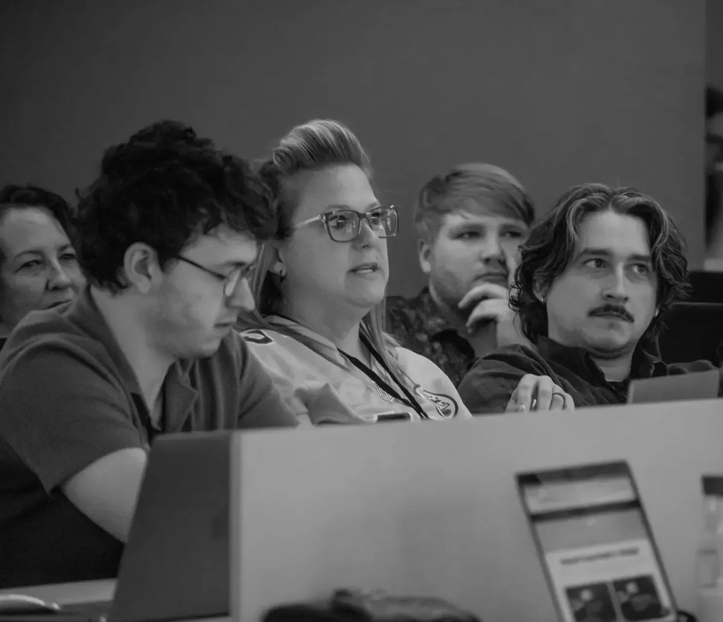 People in a classroom setting, listening attentively. Woman in glasses speaking. Other students looking on.