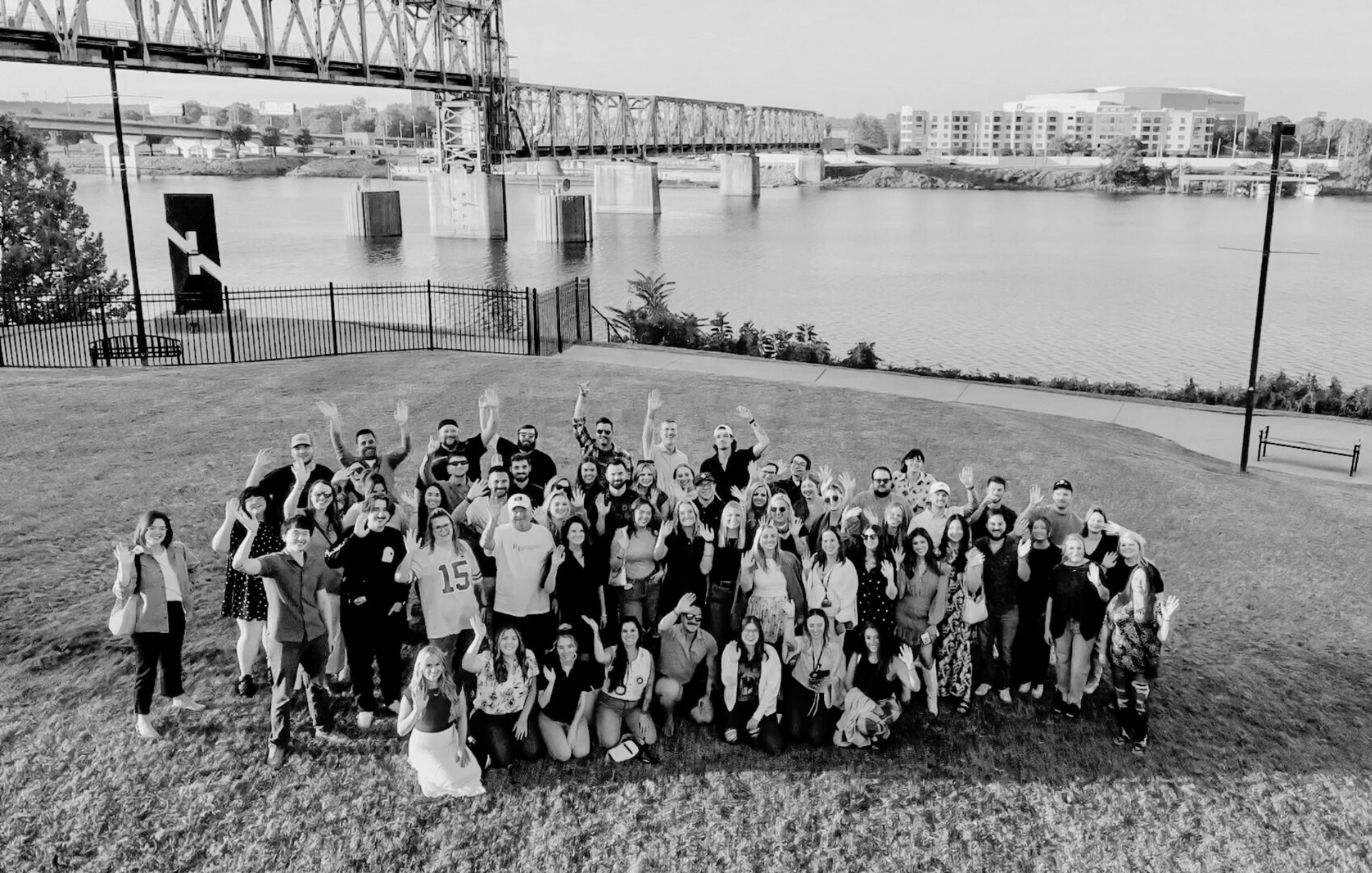 Group of people waving on a grassy hill overlooking a river and a bridge; black and white.