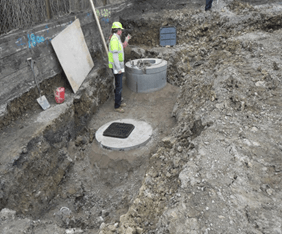 A man in a hard hat is standing next to a manhole cover in the dirt.