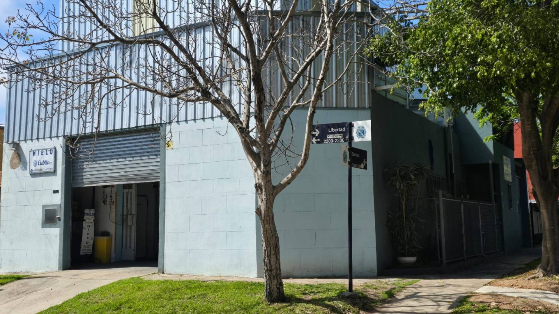 Un edificio industrial de color azul claro, situado en una esquina, con puerta enrollable y revestimiento metálico