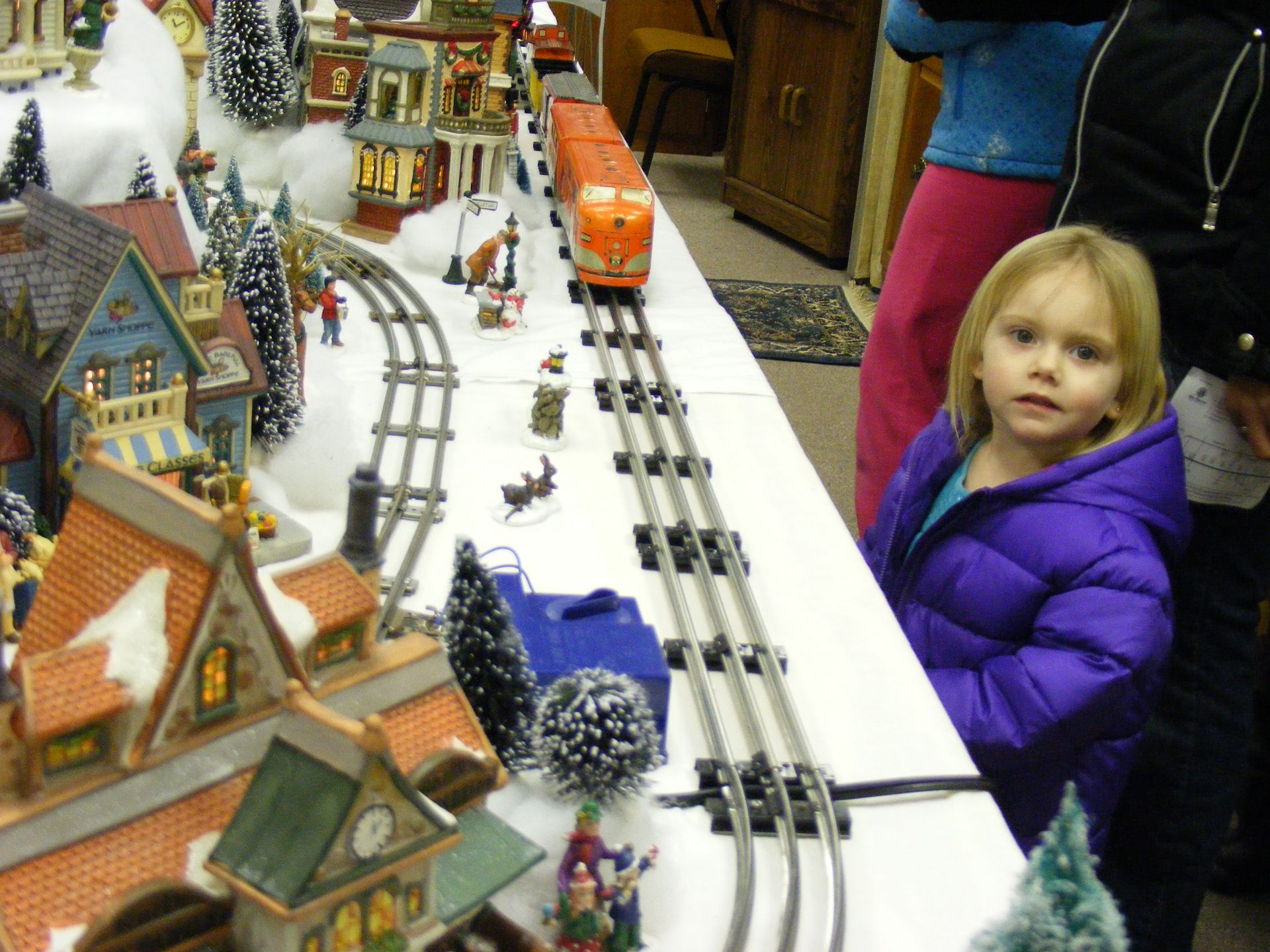 Girl watches a model train traveling through a snowy village.