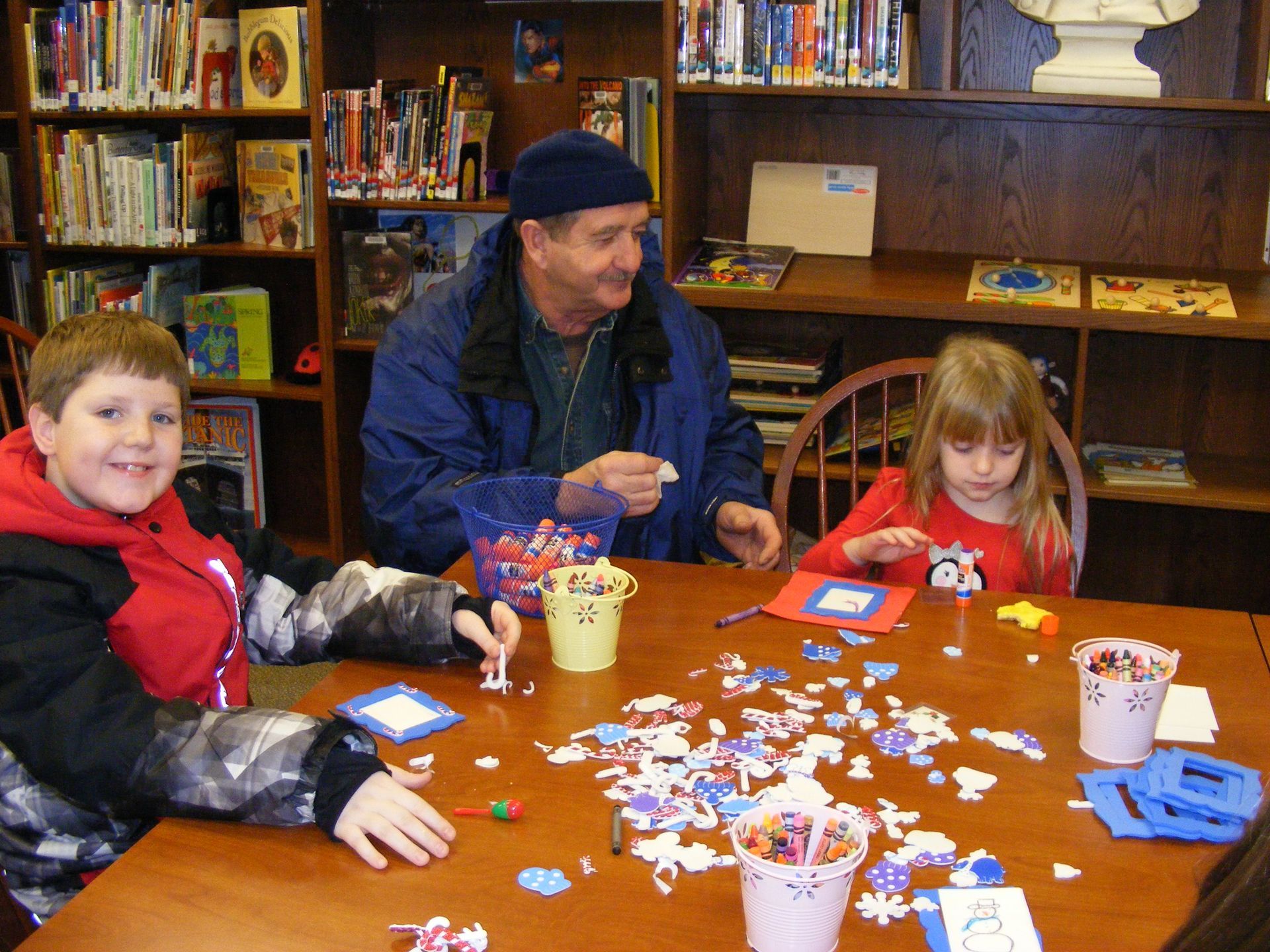 Boy, man, and girl at table in library crafting with colorful shapes.