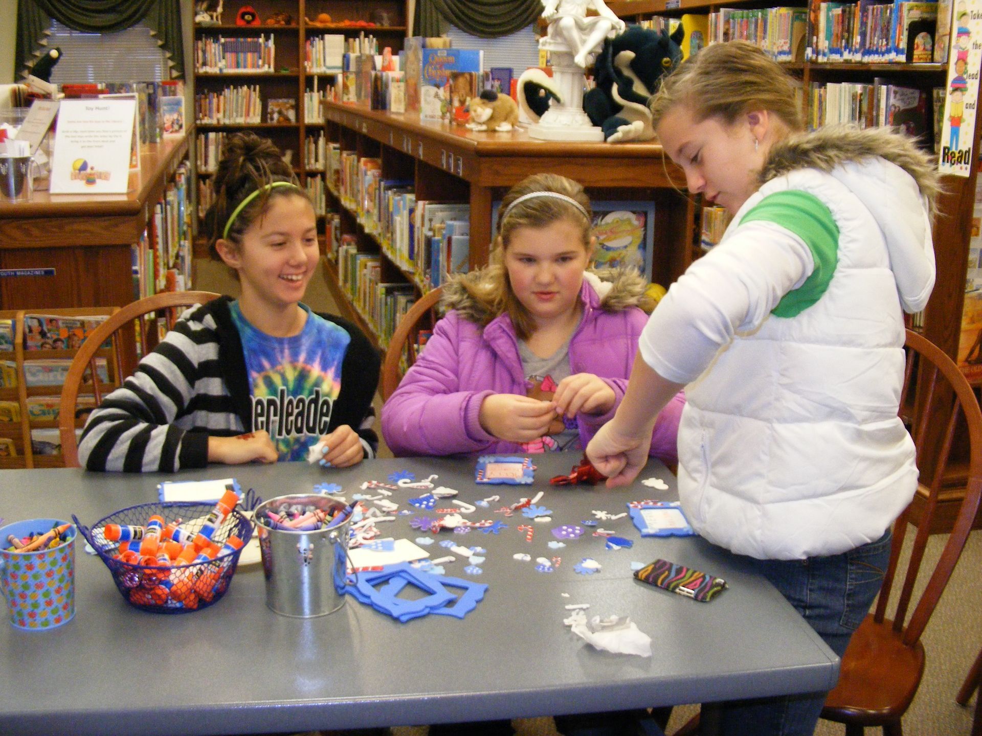 Three girls craft at a table in a library. One wears a white coat and works on a project.