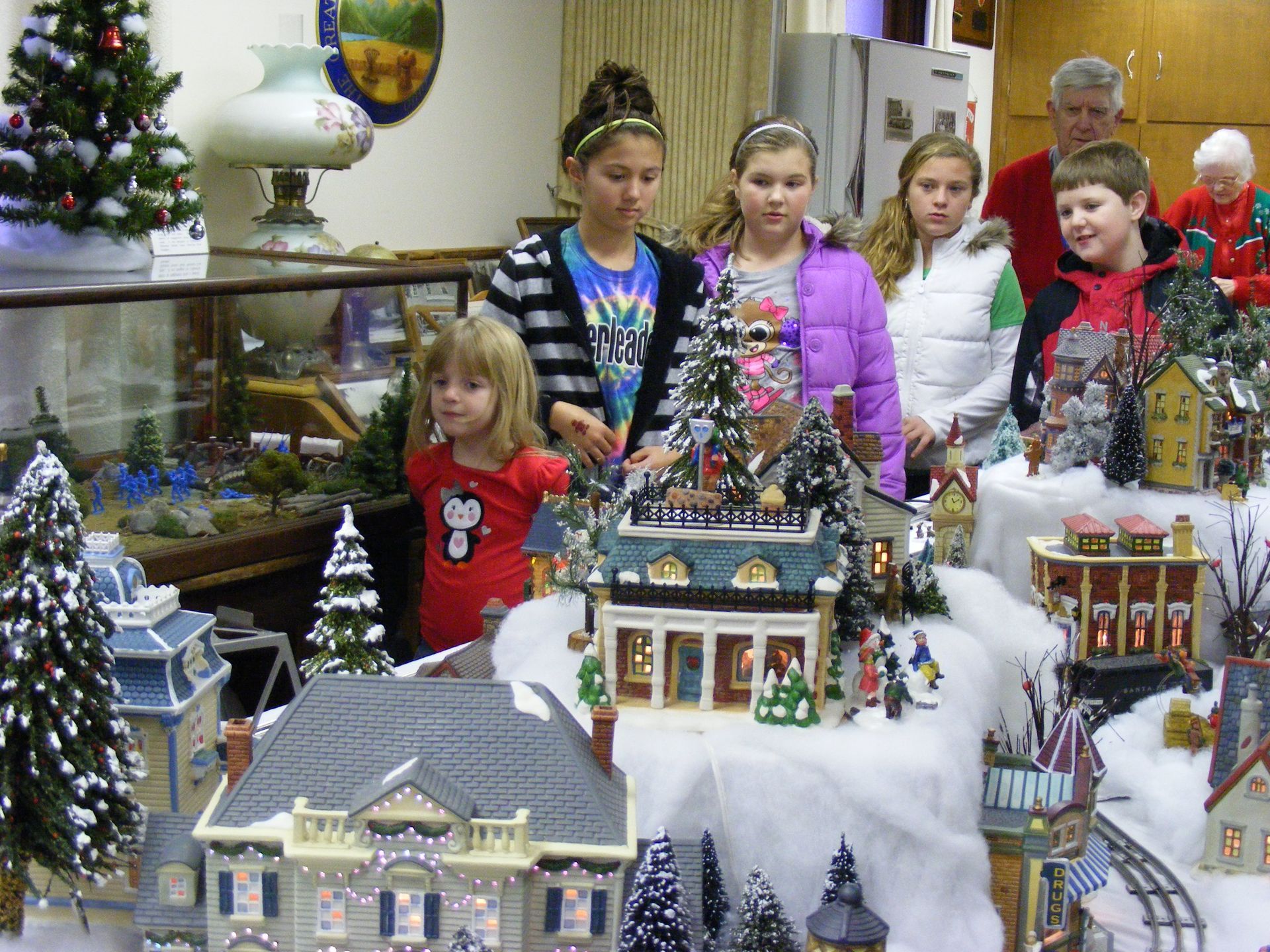 Children looking at a Christmas village display with miniature houses, trees, and figurines indoors.