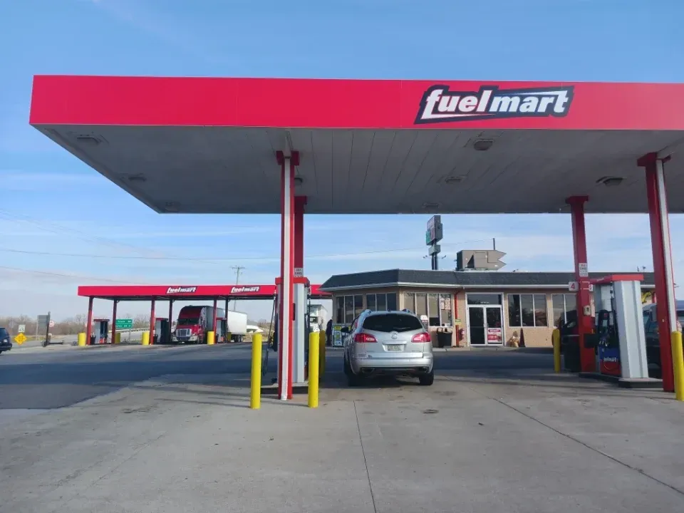 Fuelmart gas station with red canopy, vehicles, and a building, under a blue sky.