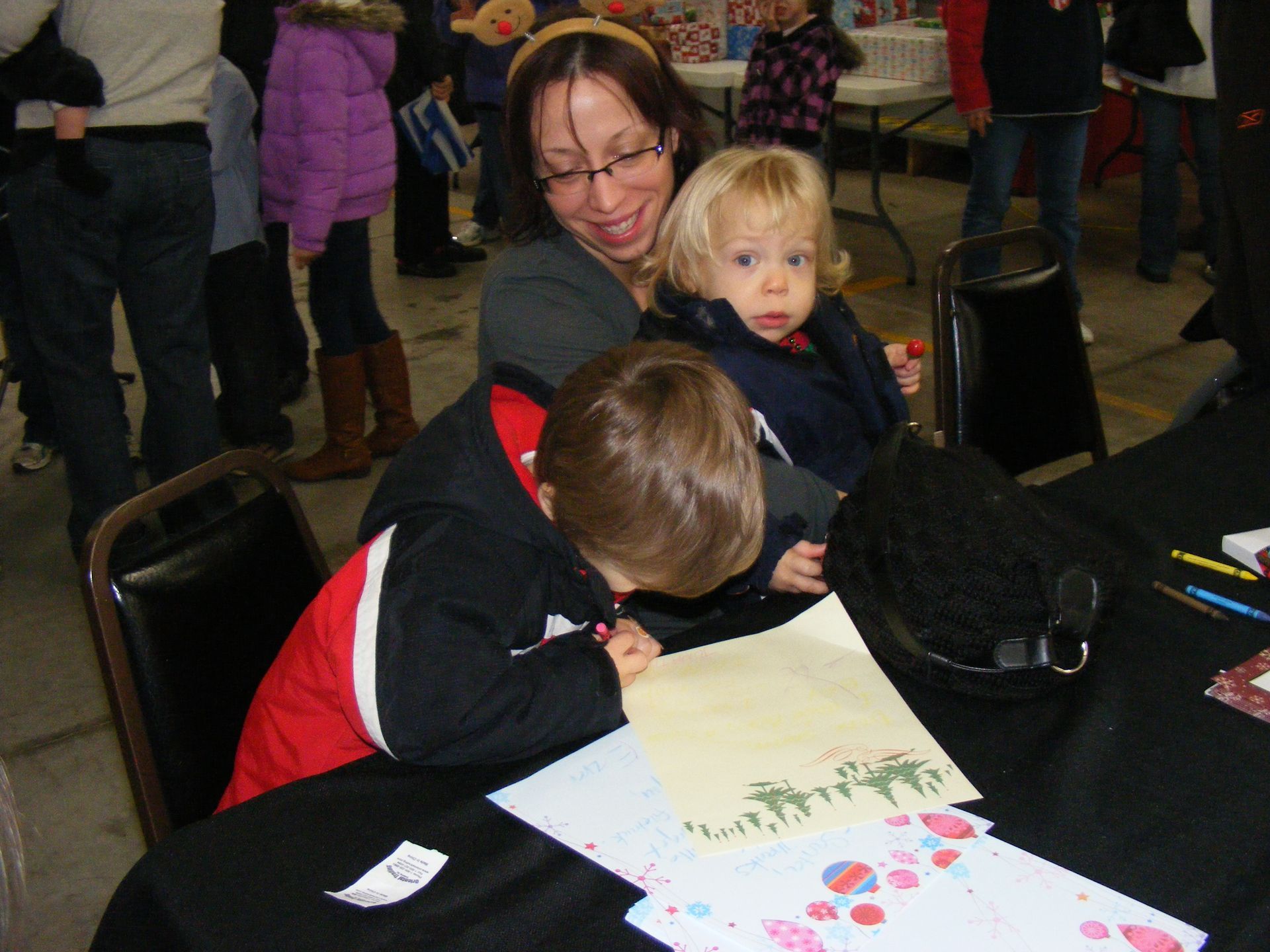 Woman with two children drawing at a table, one child drawing, other child held in lap, at an indoor event.