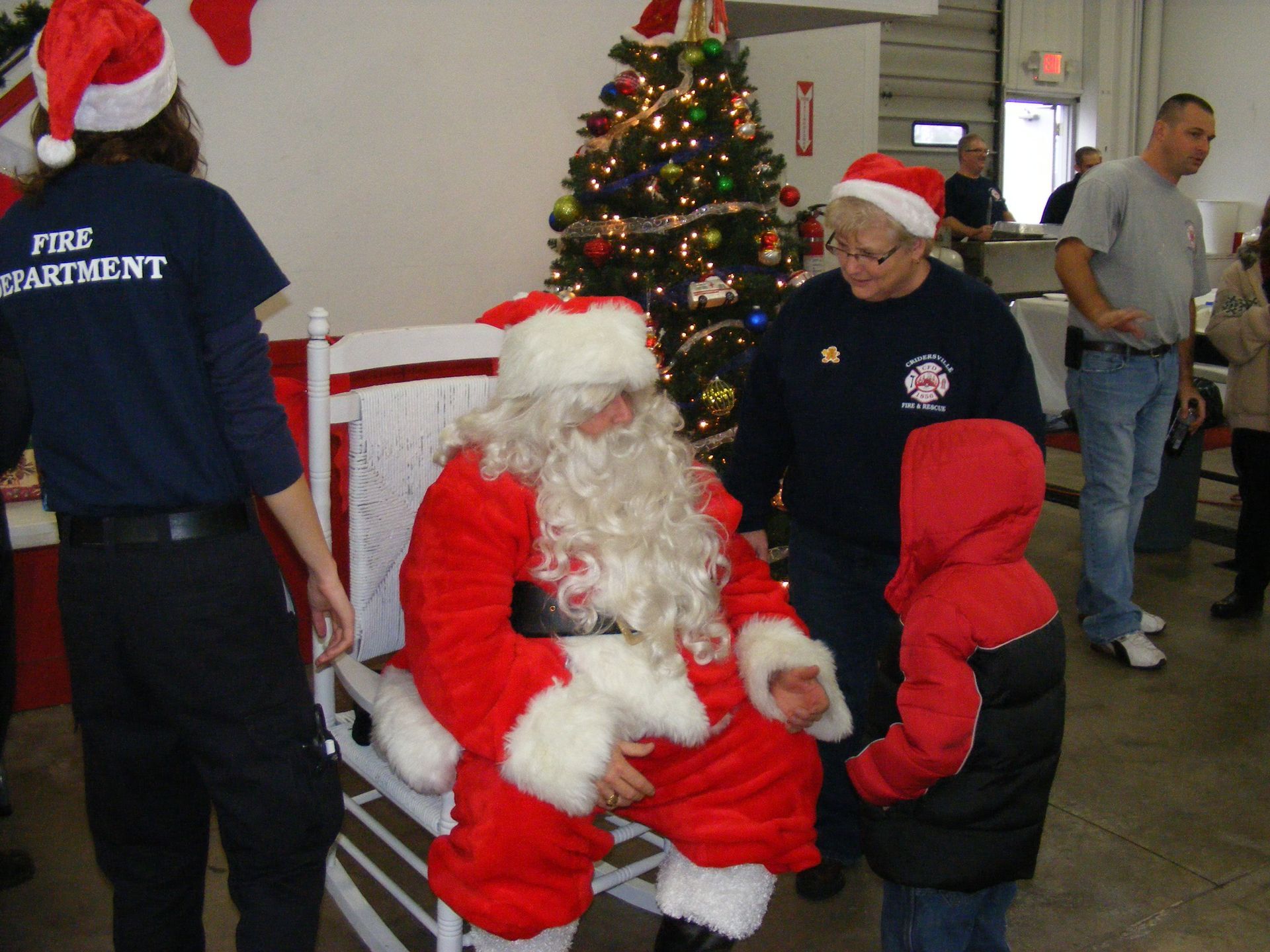 Santa Claus sits, talking to a child at a fire station Christmas event. Two firefighters nearby.