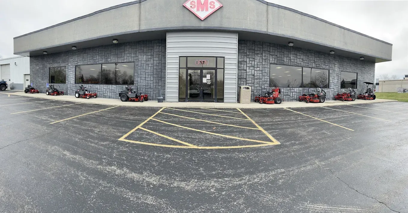 Gray stone-faced building with entrance; lawnmowers displayed outside. Cloudy day, wet parking lot.