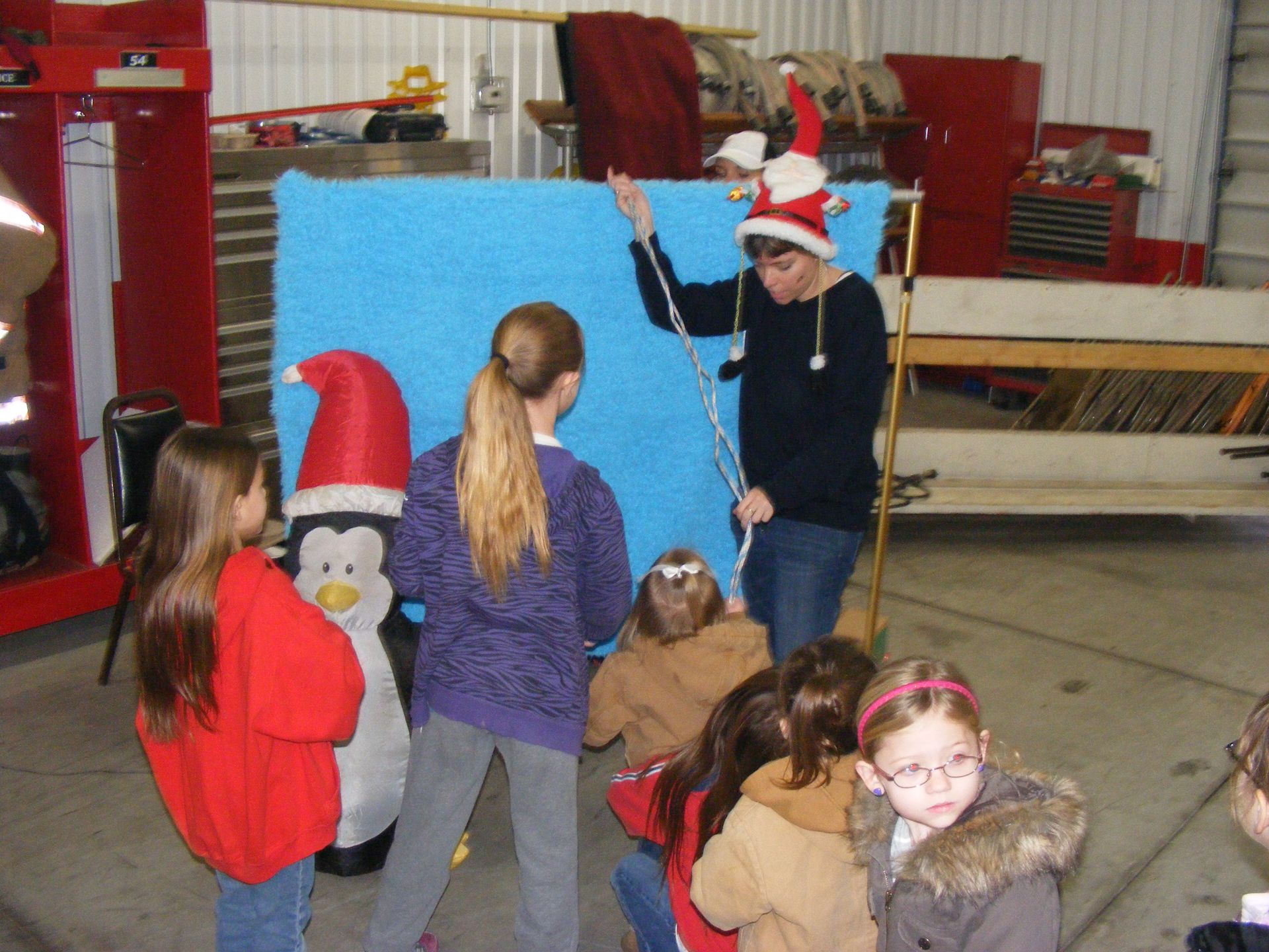 A woman in a Santa hat holds a stick in front of children, with a blue backdrop and inflatable penguin.