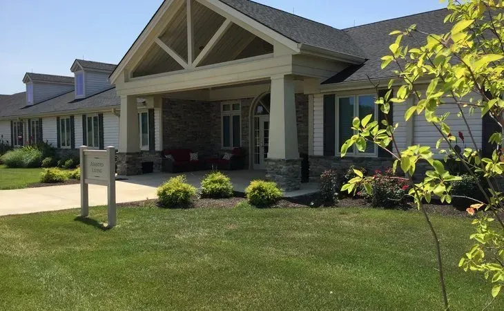Exterior view of a light-colored senior living facility with a covered porch, green lawn, and landscaping.
