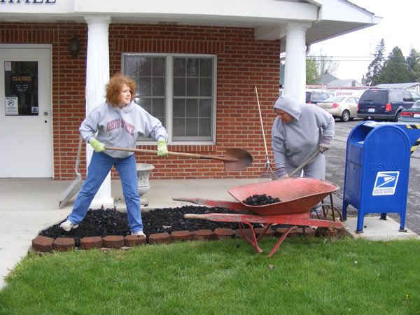 Two people spreading mulch in a flower bed in front of a building. One uses a shovel, the other a rake.