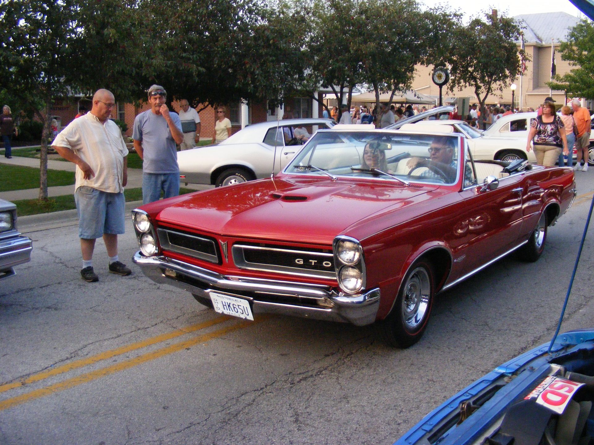 Red convertible Pontiac GTO driving on a street, car show. Two men watch. Other cars, people, and buildings are in the background.