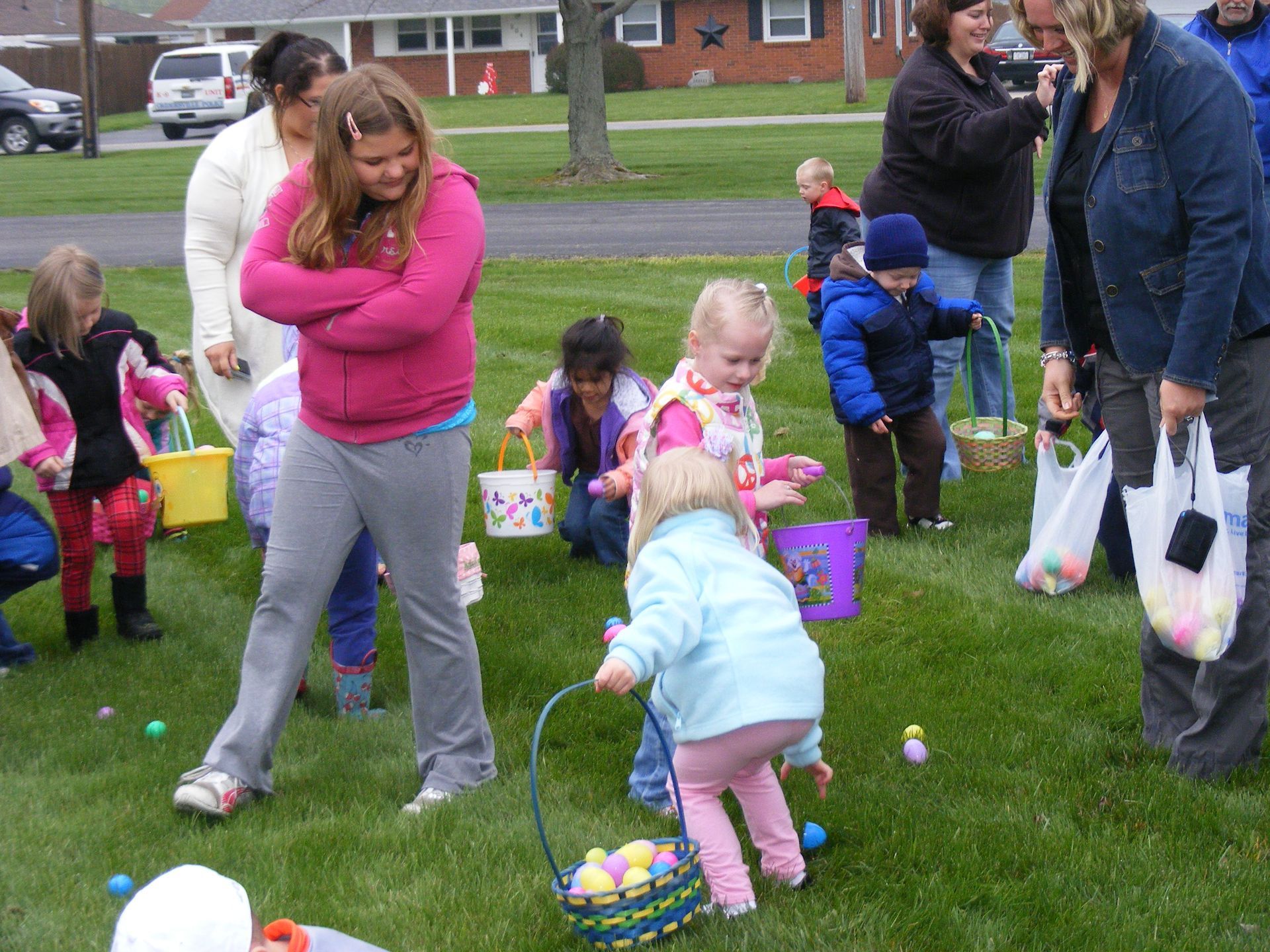 Children hunt for Easter eggs on a grassy lawn, supervised by adults.