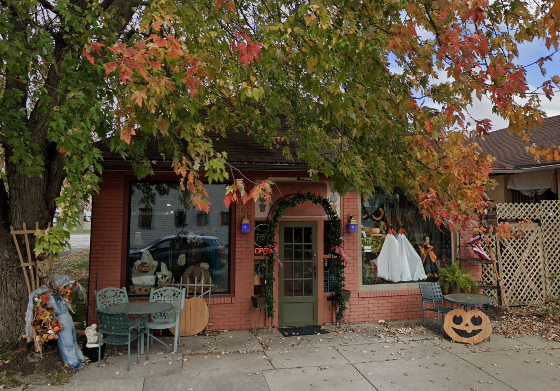 Red brick shop with fall decorations; tree with colorful leaves shades the building.