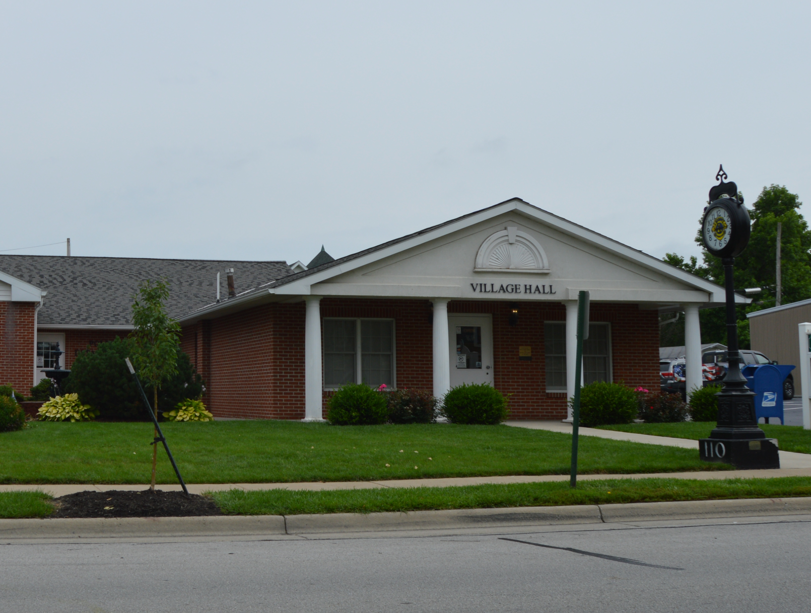 Village Hall, red brick building with a white trim, lawn, and a clock.