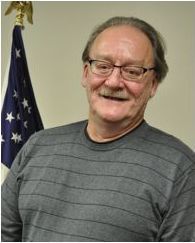 Man with glasses smiles in front of an American flag. Gray striped shirt.