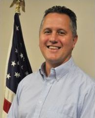 Man smiling, posing in front of an American flag. He wears a blue striped shirt.