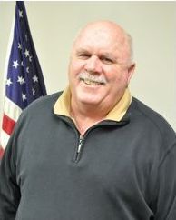 Bald man smiling, wearing a blue sweater in front of an American flag.