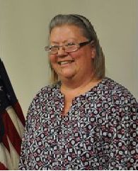 Woman with glasses smiles, stands next to US flag, wearing a patterned shirt.