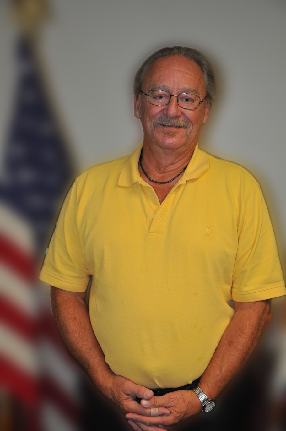 Man with glasses smiles in front of an American flag. Gray striped shirt.