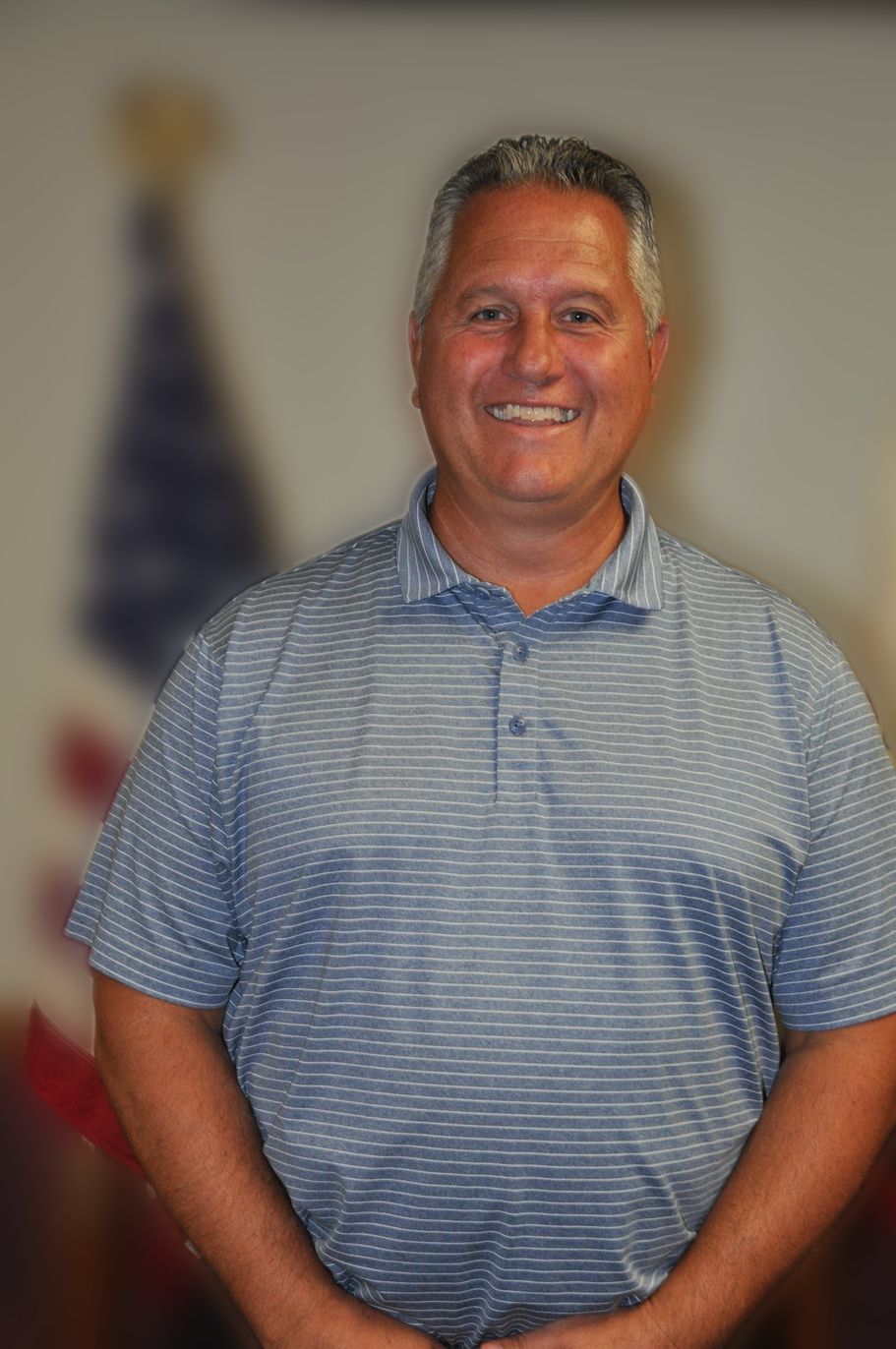 Man smiling, posing in front of an American flag. He wears a blue striped shirt.