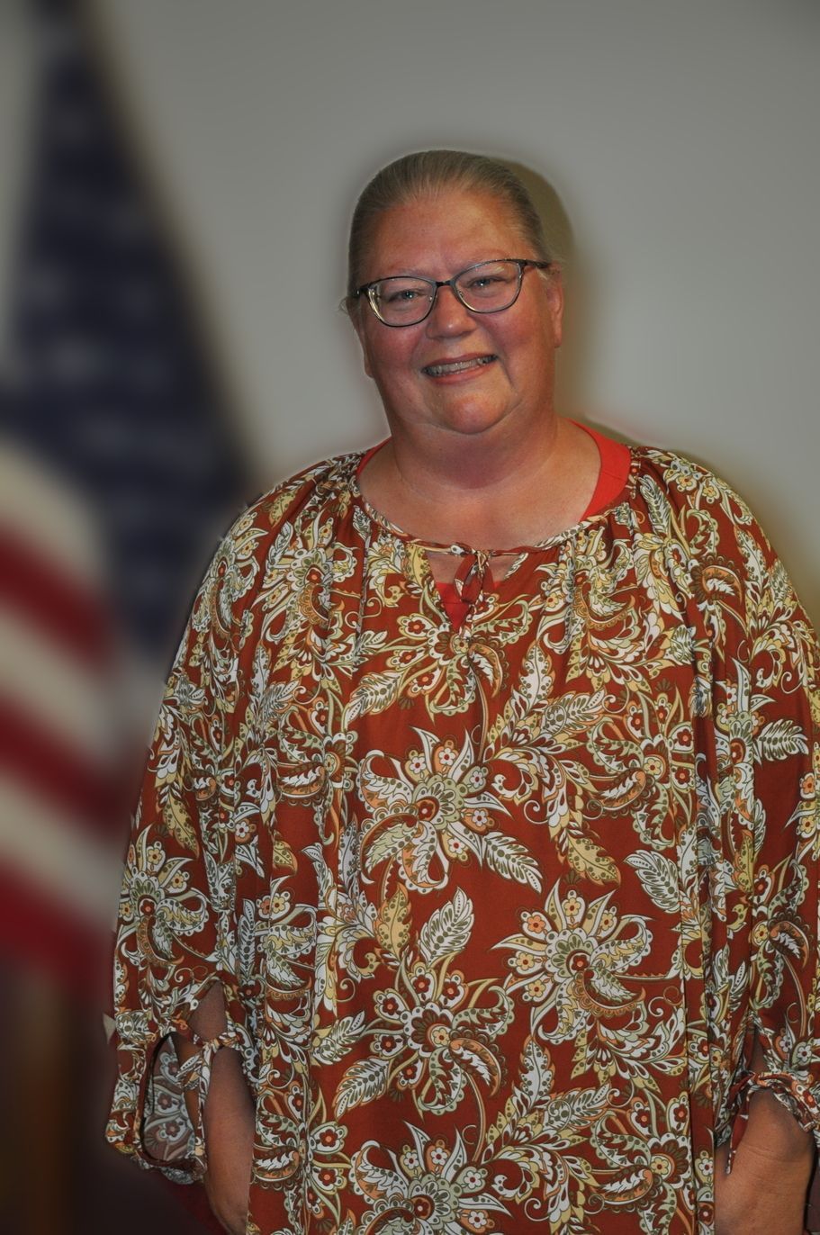 Woman with glasses smiles, stands next to US flag, wearing a patterned shirt.