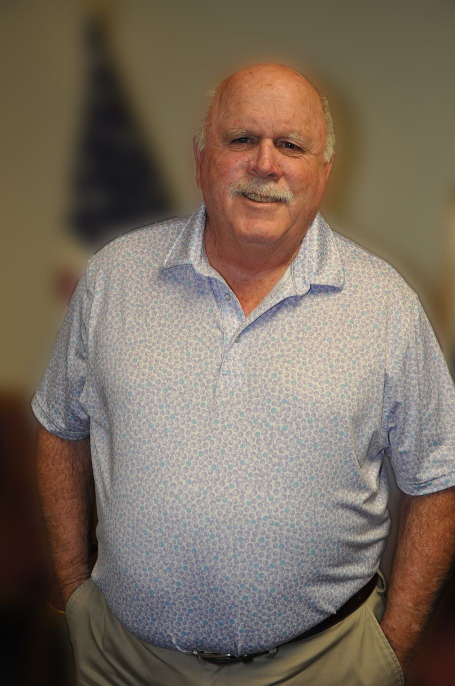 Bald man smiling, wearing a blue sweater in front of an American flag.