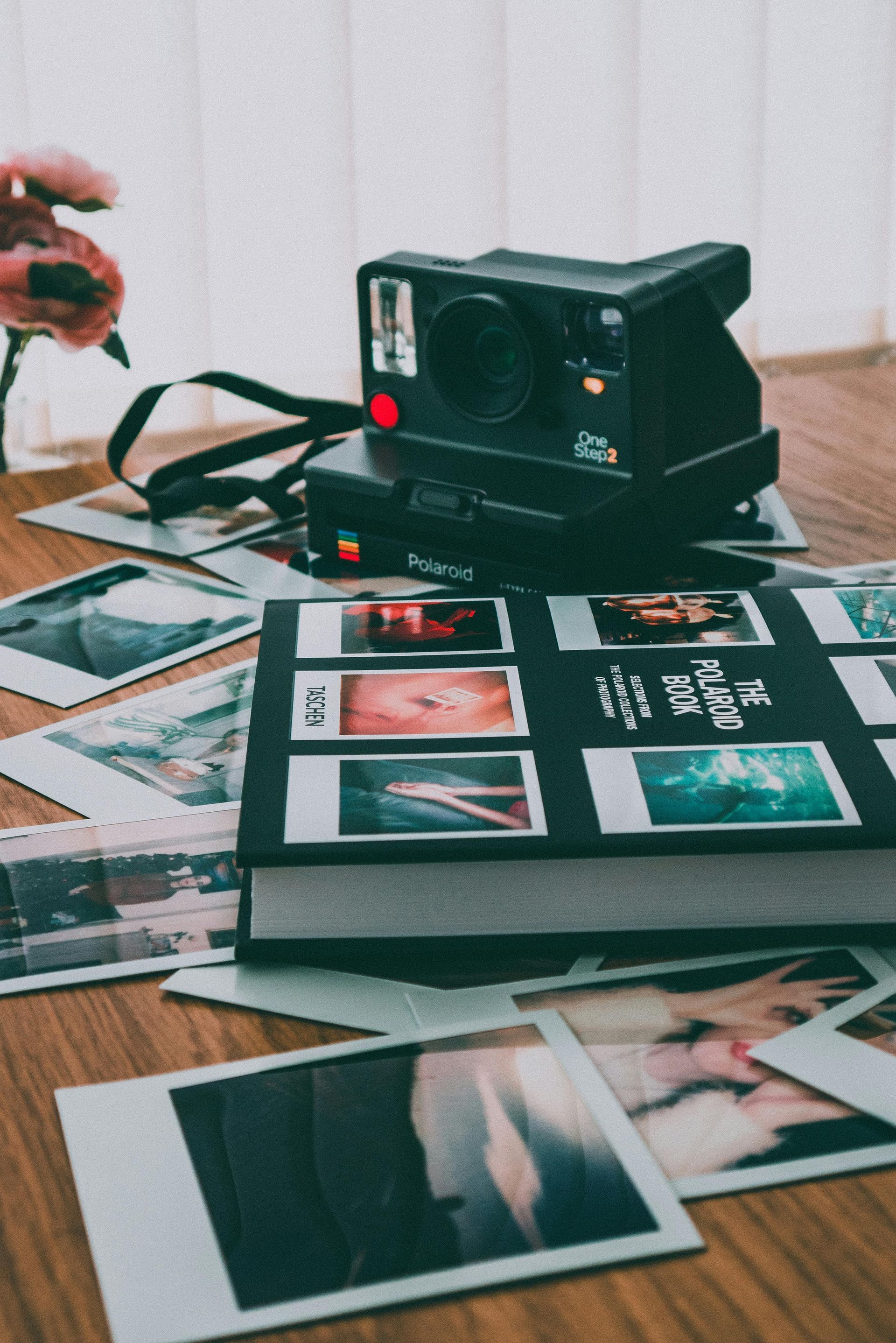 Polaroid cameras stacked on a book surrounded by printed photos on a wooden surface, near a window.