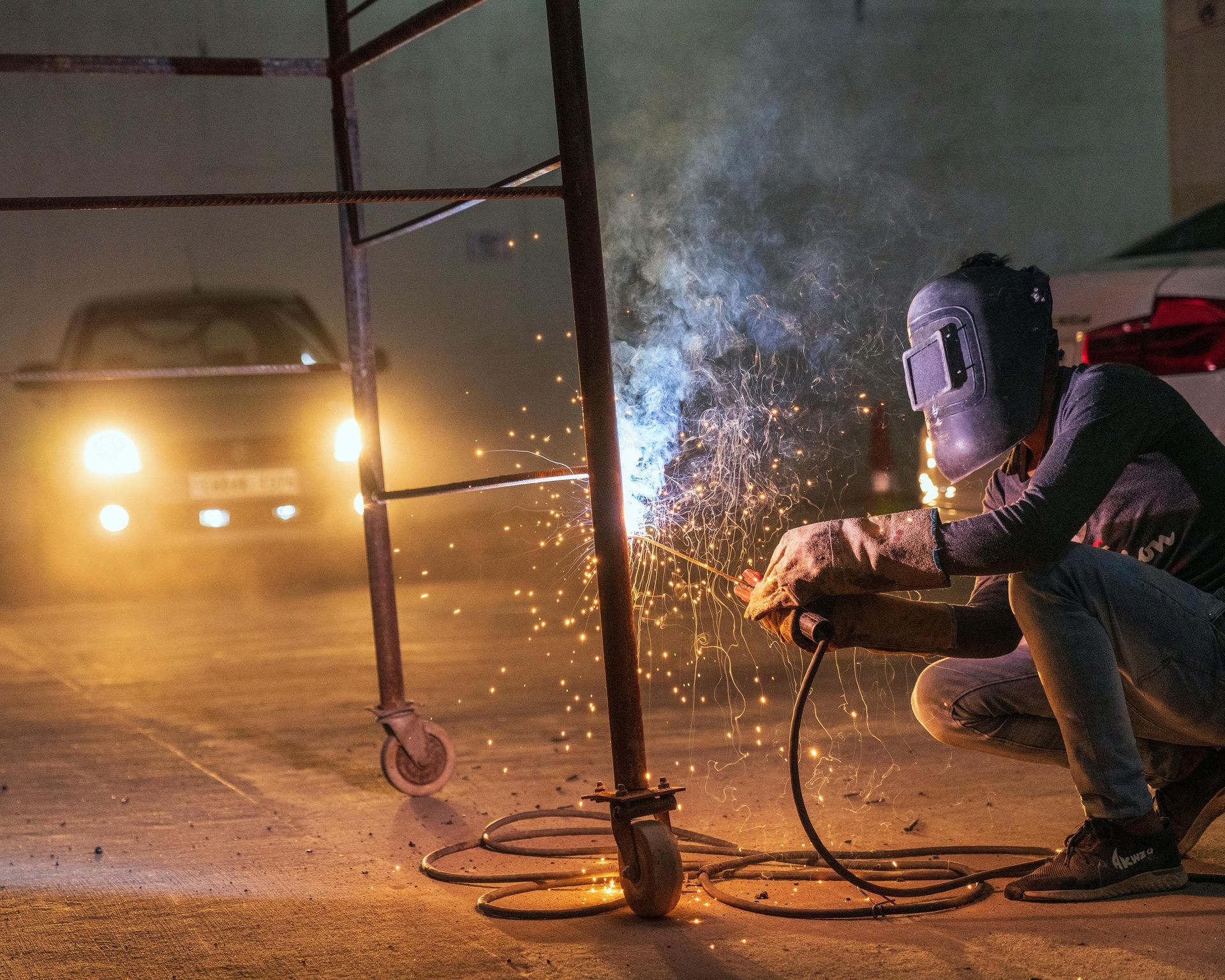 Welder in protective gear working on metal, sparks flying. Night setting with car in background.
