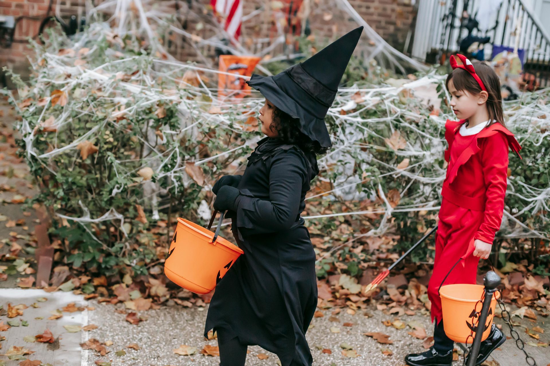 Two children in Halloween costumes trick-or-treating; a witch and a devil with orange buckets.