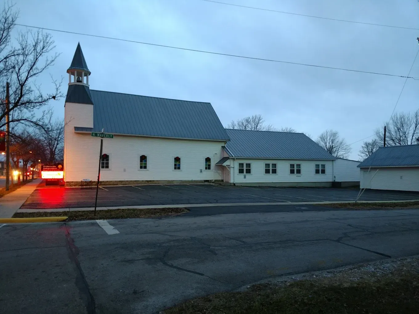 White church building with bell tower, gray roof, parking lot, and street with light at dusk.