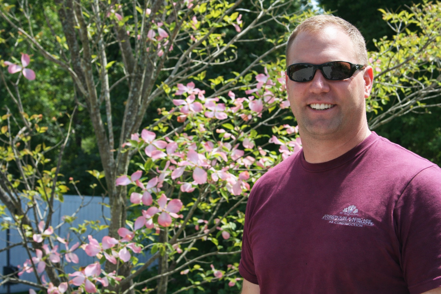 Man in sunglasses and maroon shirt standing by pink blossoms in a sunny garden