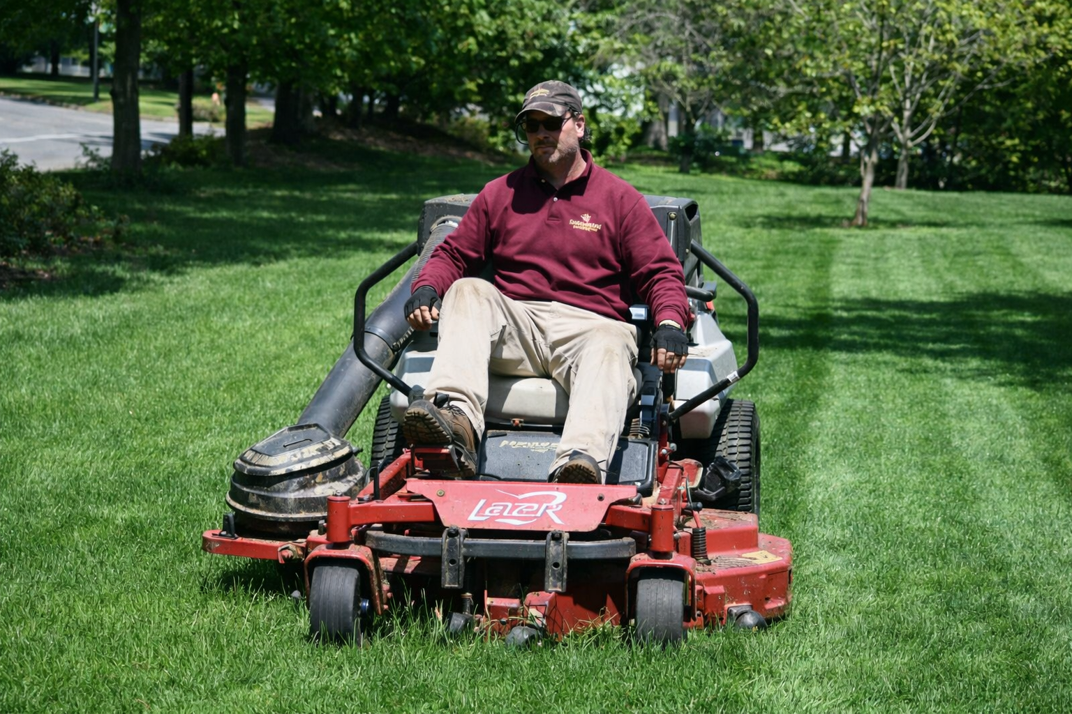 Man riding a red zero-turn lawn mower on a grassy lawn in a park