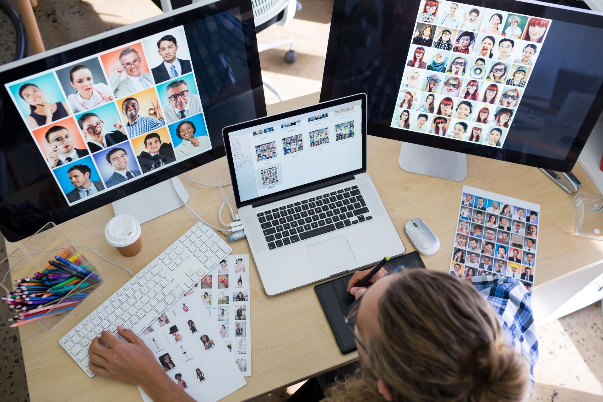 Person at desk reviewing headshot contact sheets on three computer screens and paper proofs
