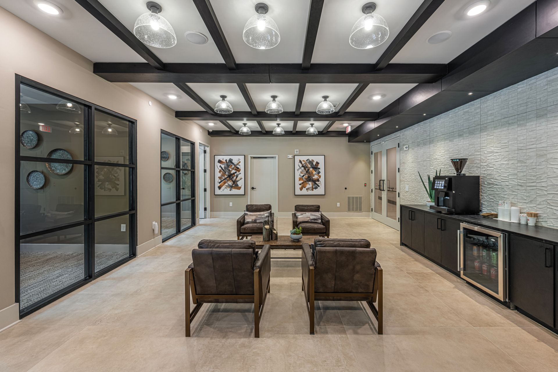 Lounge area with seating, artwork, coffee station, and glass-paneled walls. Dark beams and globe lights on the ceiling.