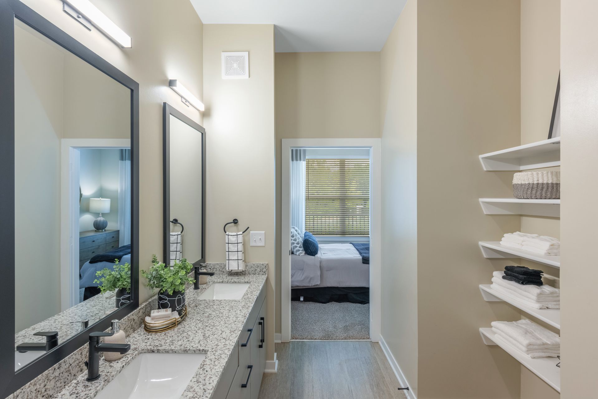 Bathroom with double vanity, mirrors, and open shelves, leading to a bedroom with bed.