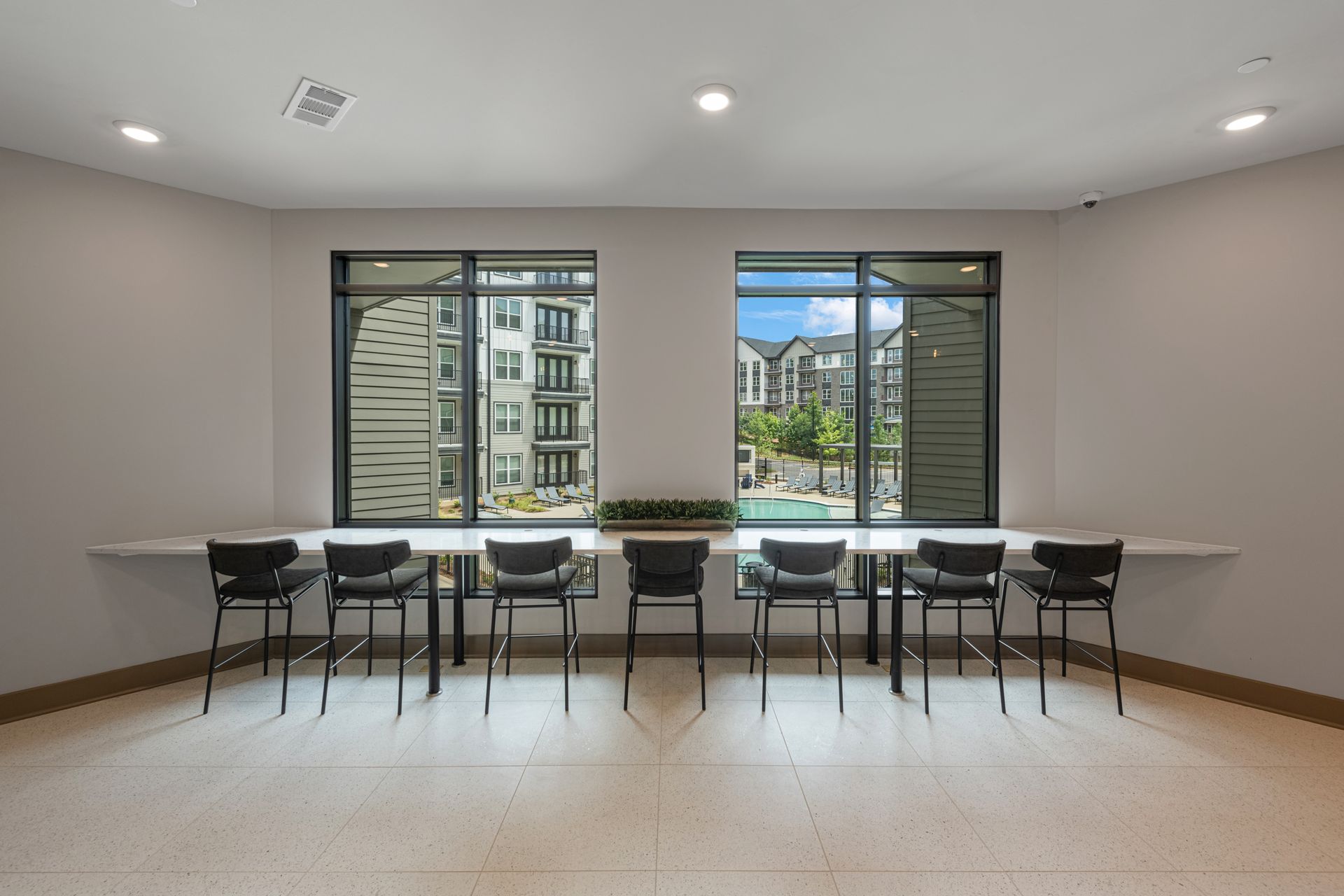 Room with large windows overlooking an apartment complex, with a long table and chairs for seating.