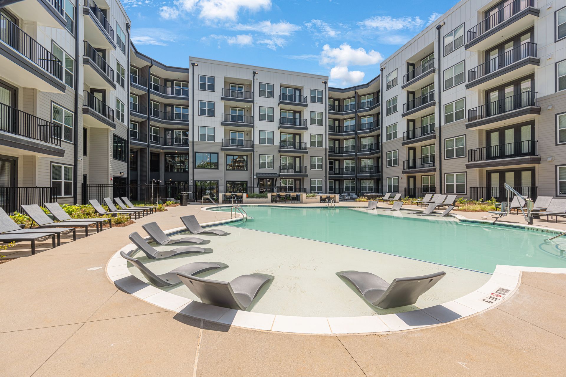 A modern apartment complex courtyard with a large swimming pool, lounge chairs on a shallow entry ledge, and a blue sky.