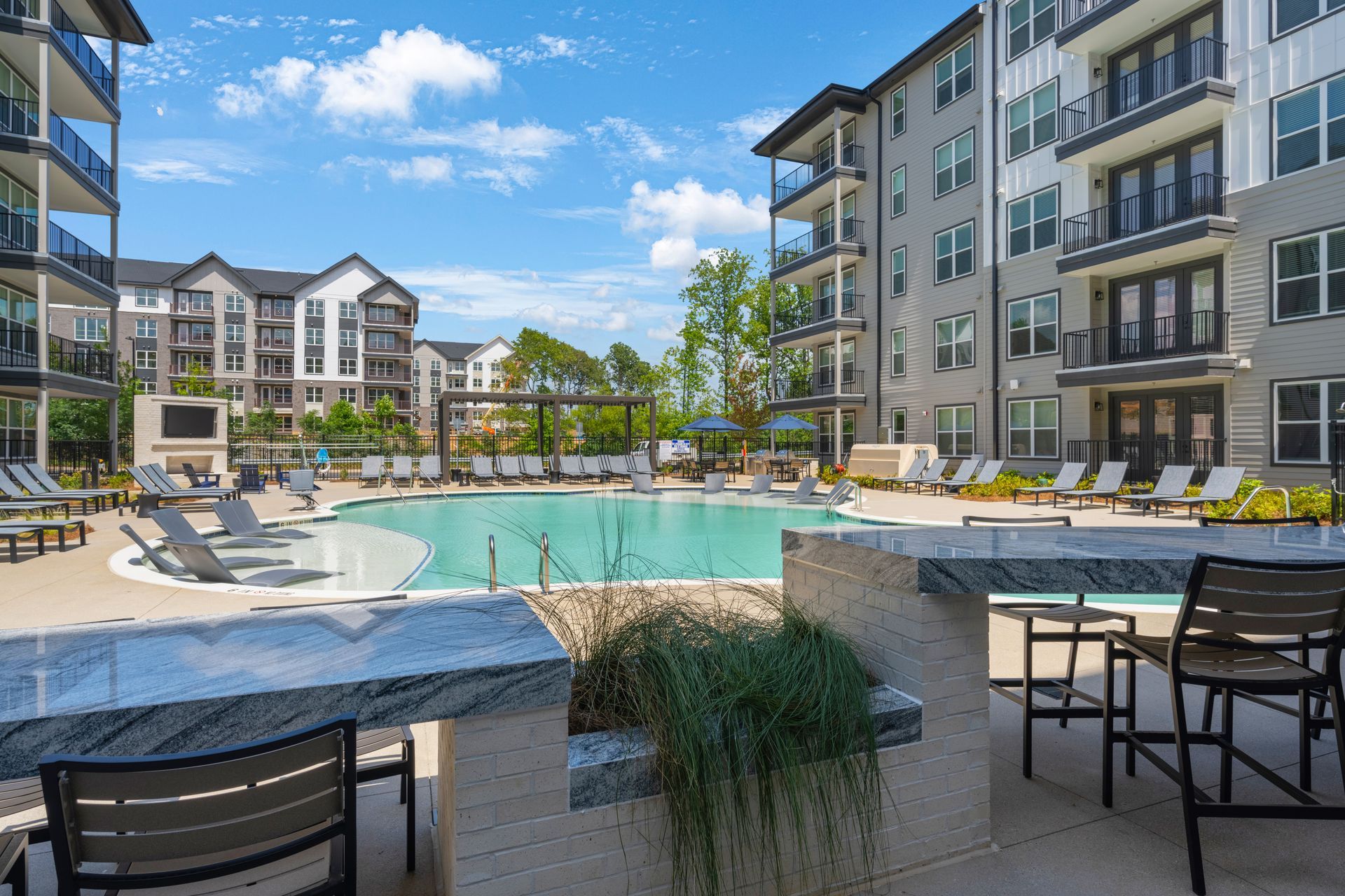 Pool area with lounge chairs, bar seating, and multi-story apartment buildings under a blue sky.