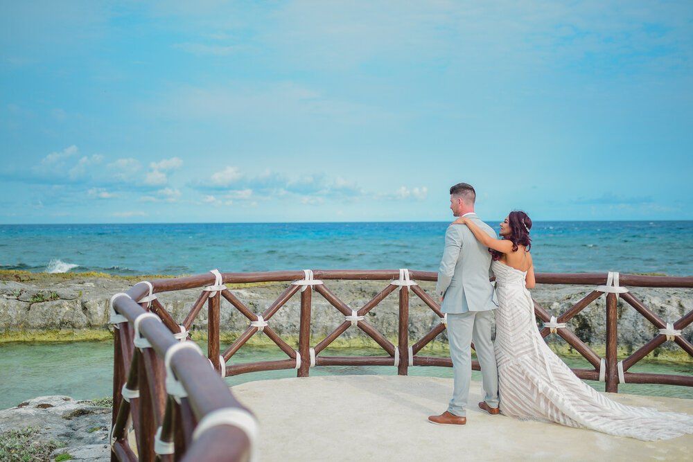 Una novia y un novio están de pie en un puente con vistas al océano.