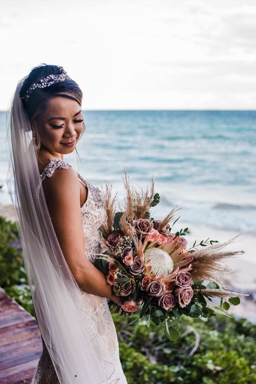 Una novia con un vestido de novia sostiene un ramo de flores frente al océano.
