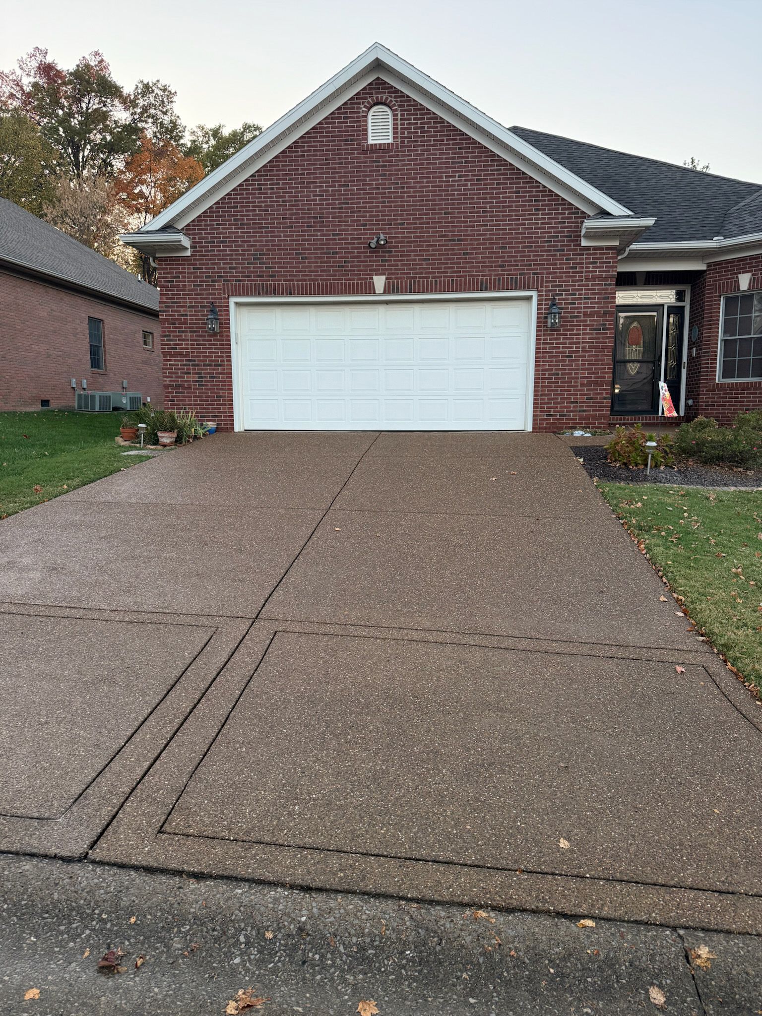 Driveway leading to a brick house with a white garage door. Trees and grass on either side.