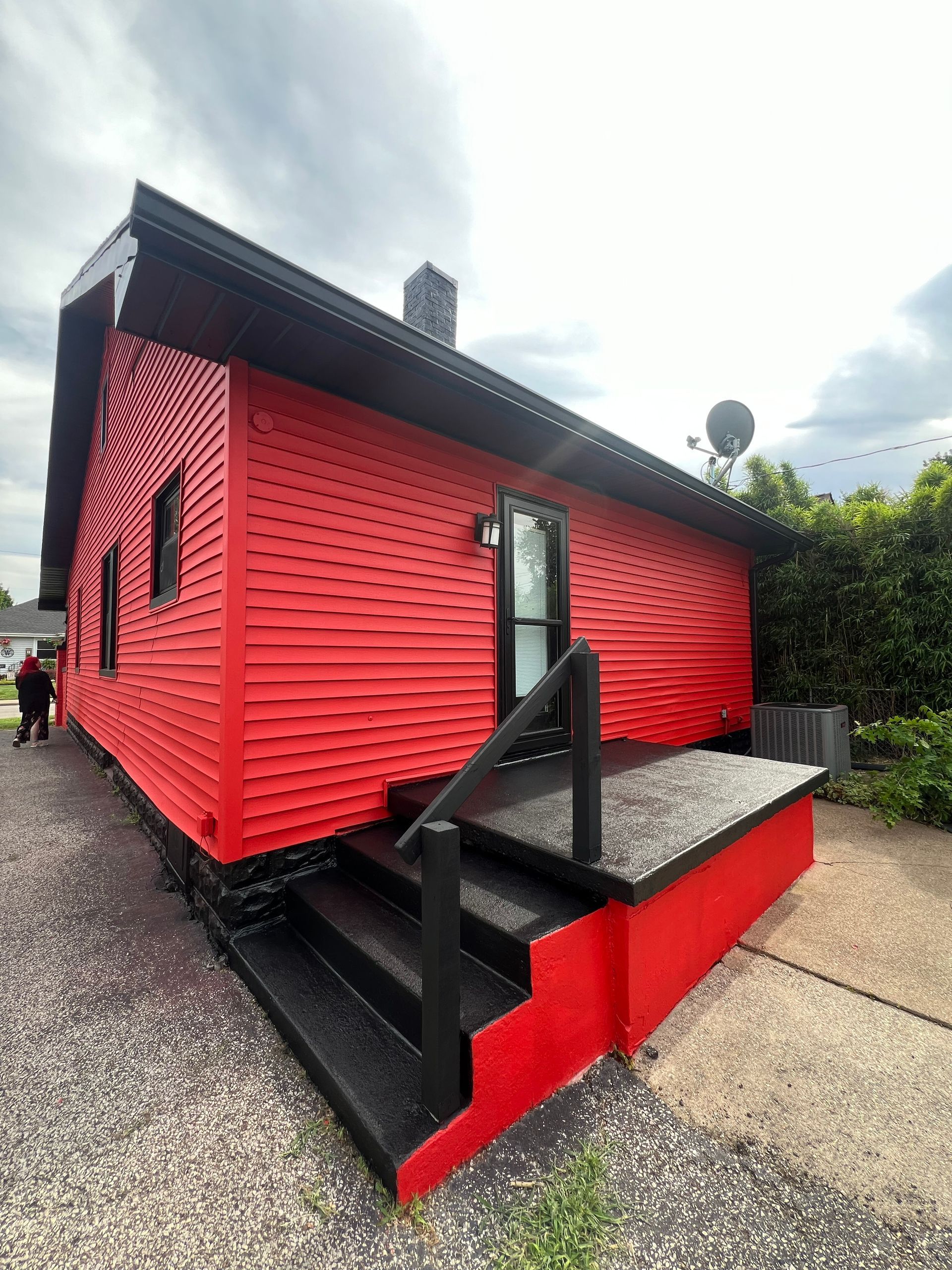 Red house with black trim, porch, and steps, on a gravel driveway, under a cloudy sky.