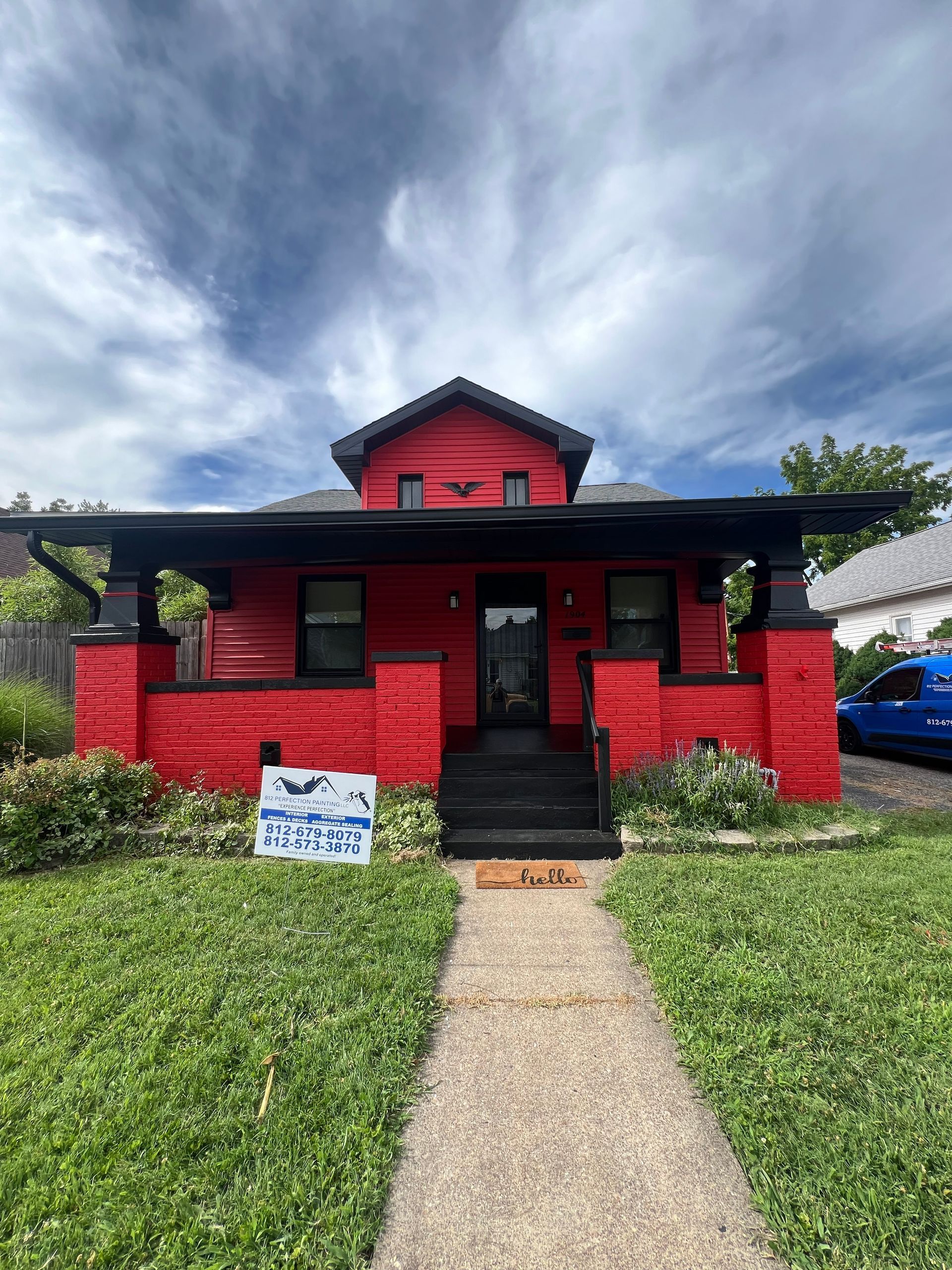 Red house with black trim and porch under a cloudy sky, with a pathway leading to the front door.