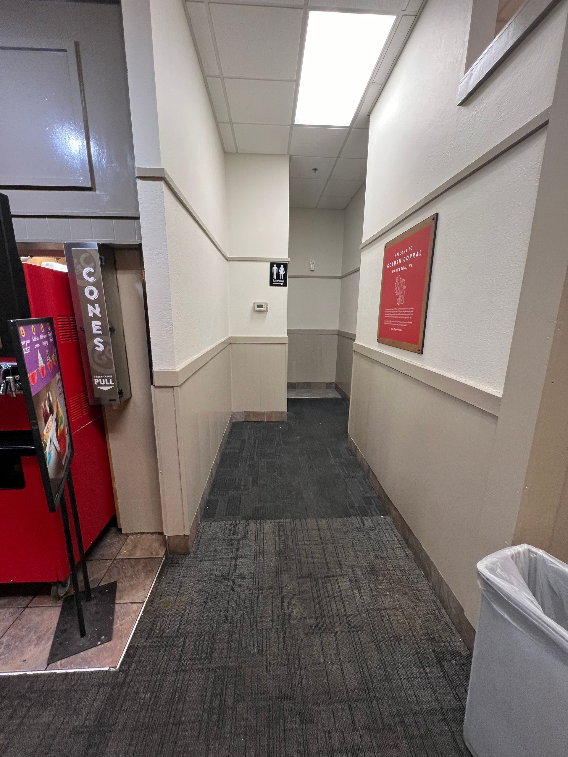 Narrow hallway with dark carpet, beige walls, a red vending machine, and a red poster.